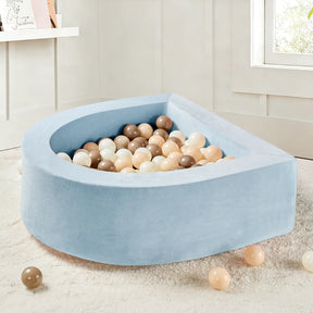 A Tiny Land® Violet PlayNest Climbing Ball Pit Set with Tunnel sits on a fluffy white rug in a bright room with white walls and a window, its light blue, semi-circular foam structure filled with beige, brown, and white balls.|Blue