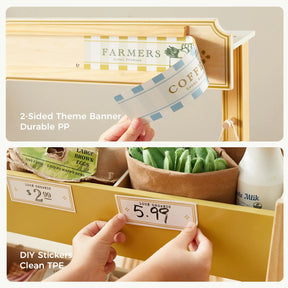 A hand attaches a Tiny Land® banner to a wooden stand and adds a $5.99 sticker to the Tiny Land® Farm-to-Table Gift Set—Light & Sound Kitchen & Market Stall, featuring 2-sided PP banner, DIY stickers, and clean TPE play food.