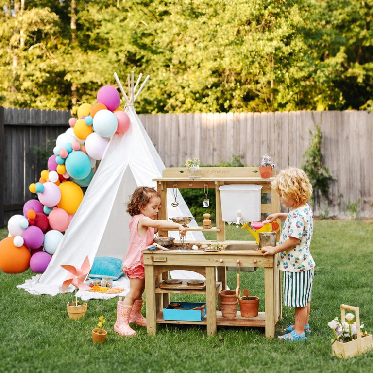 Two young children enjoy the Tiny Land® Outdoor Mud Kitchen & Sand Play Set from Tiny Land in a grassy backyard. A white teepee with colorful balloons, trees, and a wooden fence create a delightful outdoor play scene.