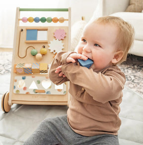 A baby in a brown shirt sits on a mat, chewing on a blue block. Behind the baby is the Tiny Land® Versatile Natural Wooden Baby Walker by Tiny Land, featuring colorful beads and shapes that invite exploration and play.