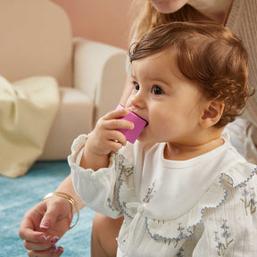 A toddler in a white outfit explores the Tiny Land Rainbow Baby Walker & Activity Cube Developmental Playset, practicing fine motor skills, while an adult sits nearby on a blue rug in a cozy room.