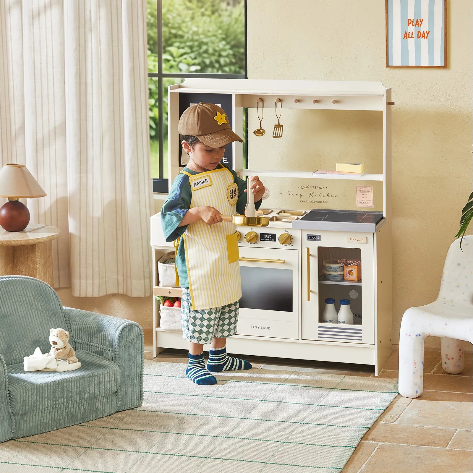 A young child wearing an apron and cap enjoys pretend play with the Tiny Land® BistroSwitch Play Kitchen, featuring toy appliances, utensils, and shelves in a cozy room. A PLAY ALL DAY sign decorates the wall while a plush chair and stuffed animal sit nearby.|White