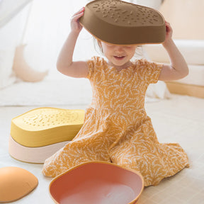 A young girl in a yellow floral dress smiles as she sits among colorful bowls, lids, and Tiny Land® Stepping Stones - Sunset Hues by Tiny Land, playfully holding a brown bowl over her head for coordination and balance fun.|Sunset Hues