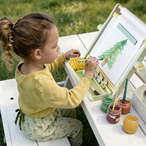 A young child sits at a white table outdoors, painting a green tree on paper with brushes and jars of paint. Dressed in a yellow top and floral pants, the child is focused on their artwork at an art station featuring the Tiny Land® Double-Sided Magnetic Tabletop Easel by Tiny Land, ready to inspire future masterpieces.|Green