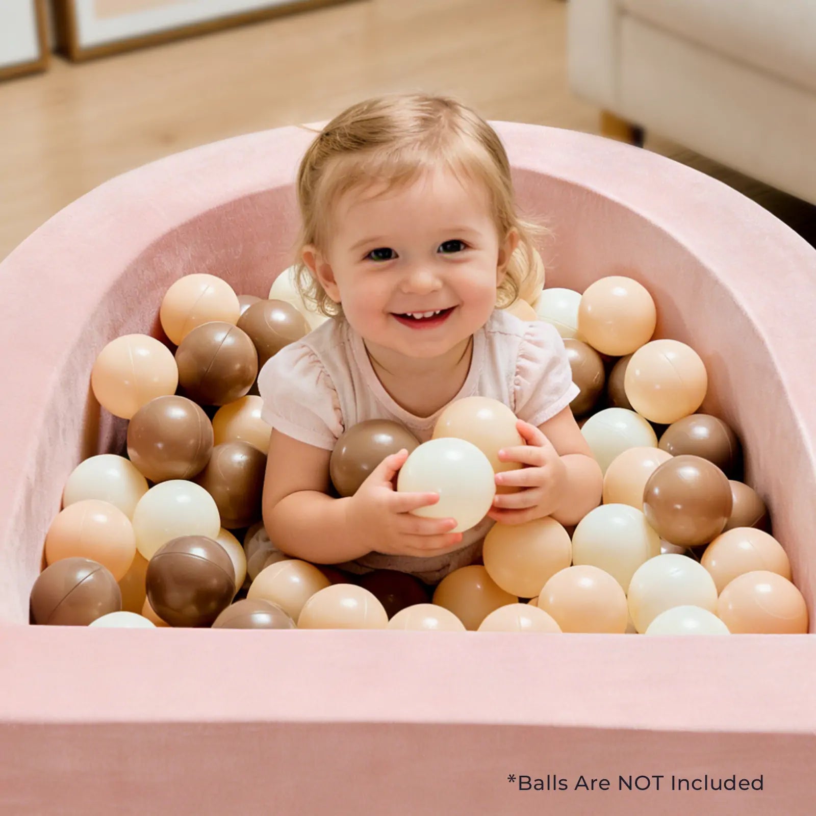 A smiling toddler sits in a Tiny Land® PlayNest Ball Pit - Ocean Ball Not Included by Tiny Land, surrounded by beige, brown, and cream balls. Text in the corner reads, *Ocean Balls Are NOT Included.|pink