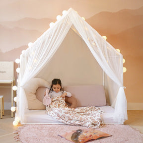 A young girl sits inside the Tiny Land® Chef’s Corner Playset—a cozy cream playhouse and kitchen set—surrounded by soft pillows and a pink rug, with string lights above, reading a book under a patterned blanket.|mesh