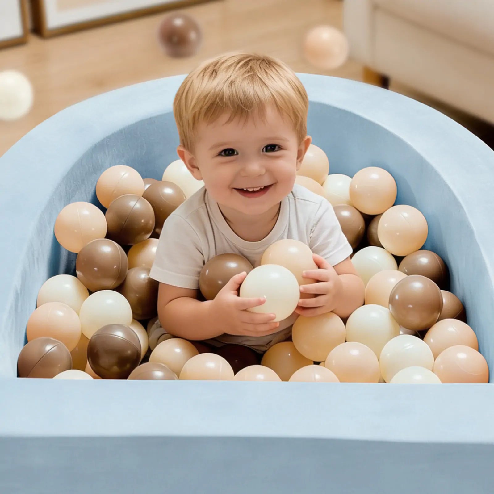 A young child smiles while sitting in the Tiny Land® PlayNest Ball Pit with 200 beige, brown, and cream ocean balls by Tiny Land, surrounded by a cozy blue high-density foam enclosure.