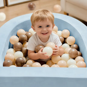A young child smiles while sitting in the Tiny Land® PlayNest Ball Pit with 200 beige, brown, and cream ocean balls by Tiny Land, surrounded by a cozy blue high-density foam enclosure.