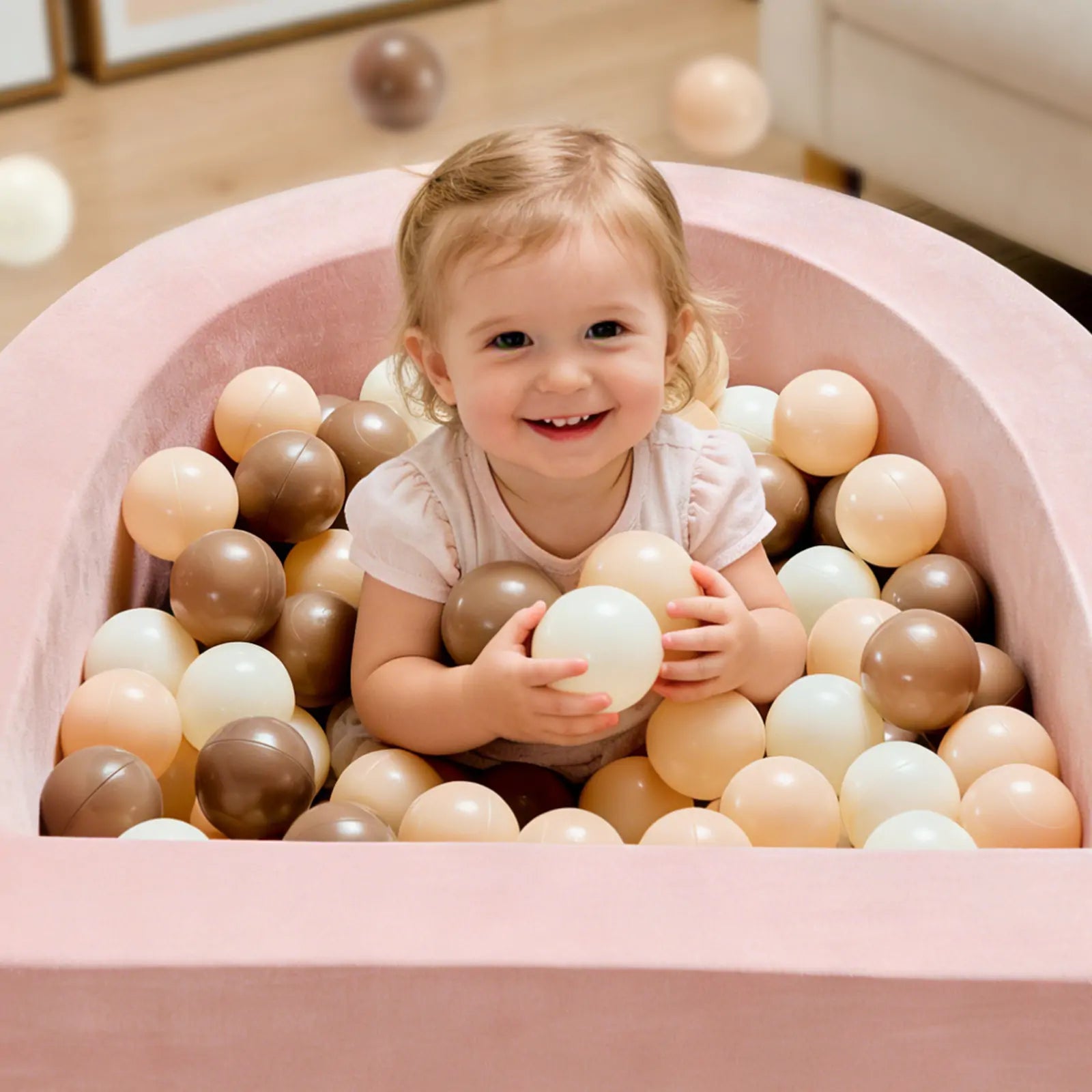 A smiling toddler sits in the Tiny Land® PlayNest Ball Pit with 200 beige, white, and brown ocean balls by Tiny Land, holding a white ball and looking up. Some balls are scattered on the wooden floor in this cozy play space.