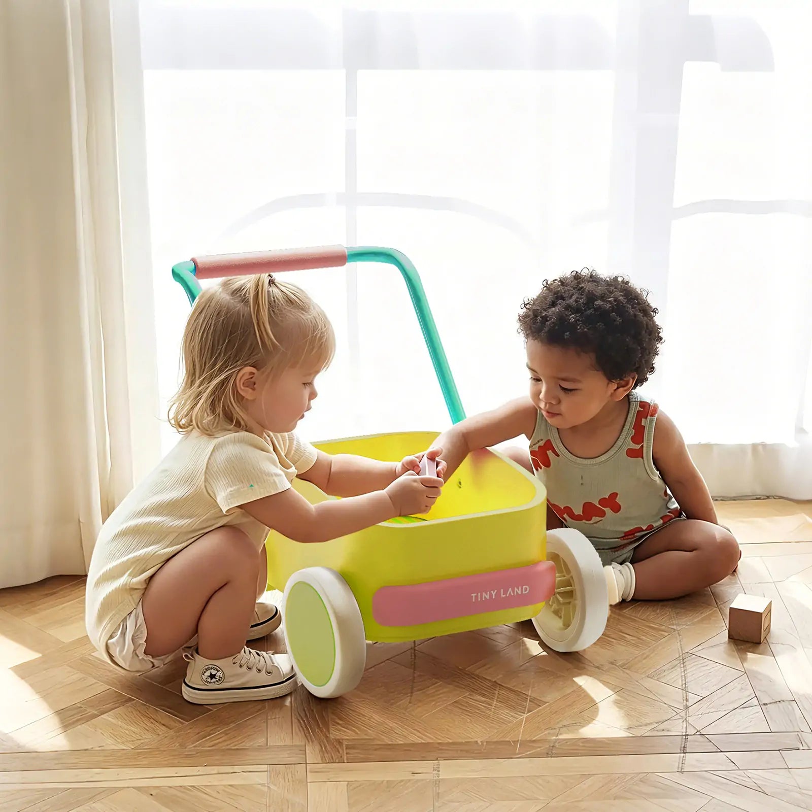 Two young children play on a wooden floor with the Tiny Land® 3-in-1 Wooden Baby Walker Wagon - Spring Color. Sunlight streams through sheer curtains. One child wears light clothes, the other sports a gray tank top with orange shapes.