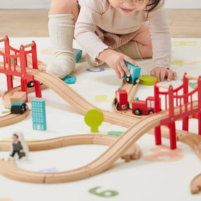 A young child kneels on a mat, wearing light clothing and socks, playing with the Tiny Land® Wooden Train Set for Children 39 Pcs by Tiny Land. The eco-friendly set includes curved tracks, red bridges, buildings, trees, and a blue train car.|39 Pcs
