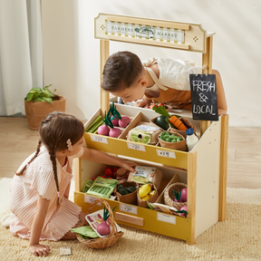 Two young children play at a wooden farmer’s market stand with the Tiny Land Play Food Organic Play Food Collection, featuring realistic toddler play food, labeled bins, a "Fresh & Local" sign, and baskets on a light indoor rug.