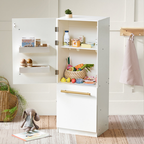 A white toy refrigerator stands open, showing play food like fruits, veggies, eggs, and milk. Nearby is a Tiny Land Play Food Organic Meat & Seafood Pack beside a stuffed rabbit on the rug, with a woven basket in the background.