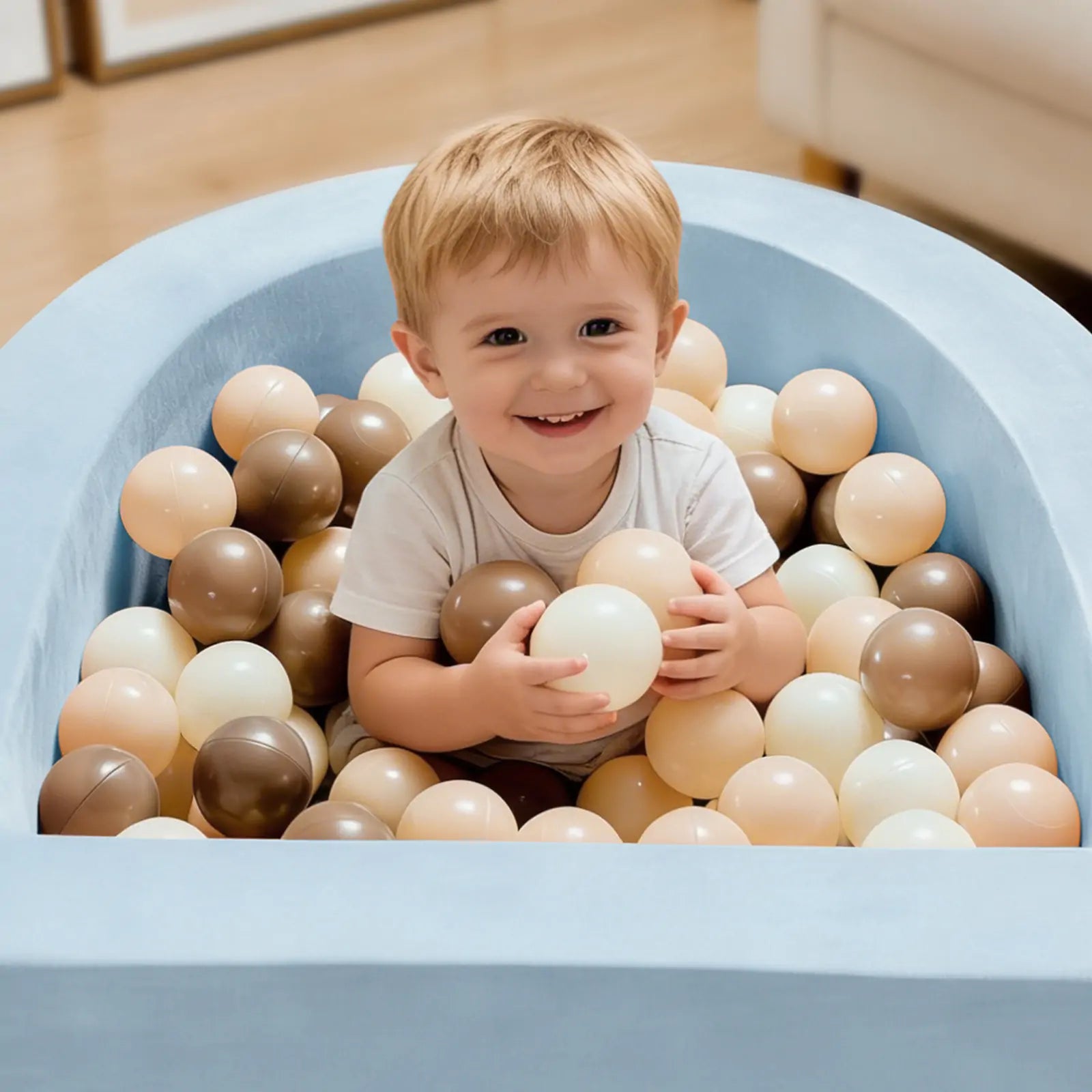 A smiling toddler sits in the Tiny Land® PlayNest Climbing Ball Pit Set with Tunnel by Tiny Land—a light blue, curved foam ball pit filled with beige, brown, and white plastic balls—holding one ball while looking up.|Blue