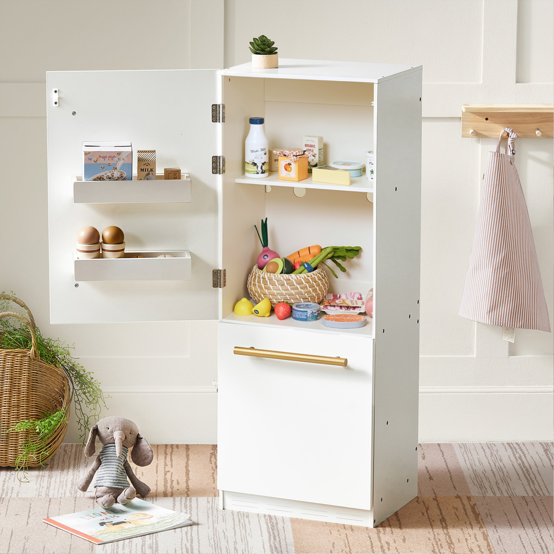 A white toy refrigerator stands open, displaying the Tiny Land® Egg Combo Playset from Tiny Land Play Food with realistic eggs inside. Nearby, a plush bunny rests on a rug, and a wicker basket and apron hang in the background.