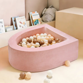 The Tiny Land® PlayNest Ball Pit with 200 Ocean Balls by Tiny Land, a soft pink teardrop-shaped foam pit filled with pastel balls, sits in a cozy, modern toddler play space with books and a stuffed animal in the background.