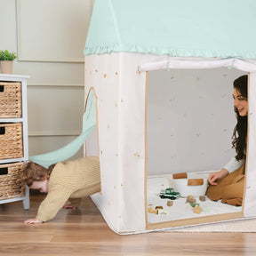 A toddler crawls out of the Tiny Land® Sweet Woodland Kids Play Tent while a woman smiles inside. Wooden toys and blocks rest on the included play mat, and a small wicker drawer unit from Tiny Land is nearby.|green