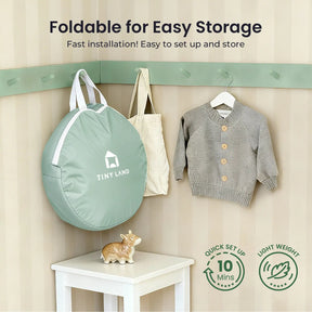 A sage green Tiny Land® Compact Adventure 3-in-1 Play Tunnel (no balls) hangs on a wall hook next to a child’s gray sweater, with a decorative hippo below. Lightweight and easy to set up—perfect for storing your play tunnel.|green
