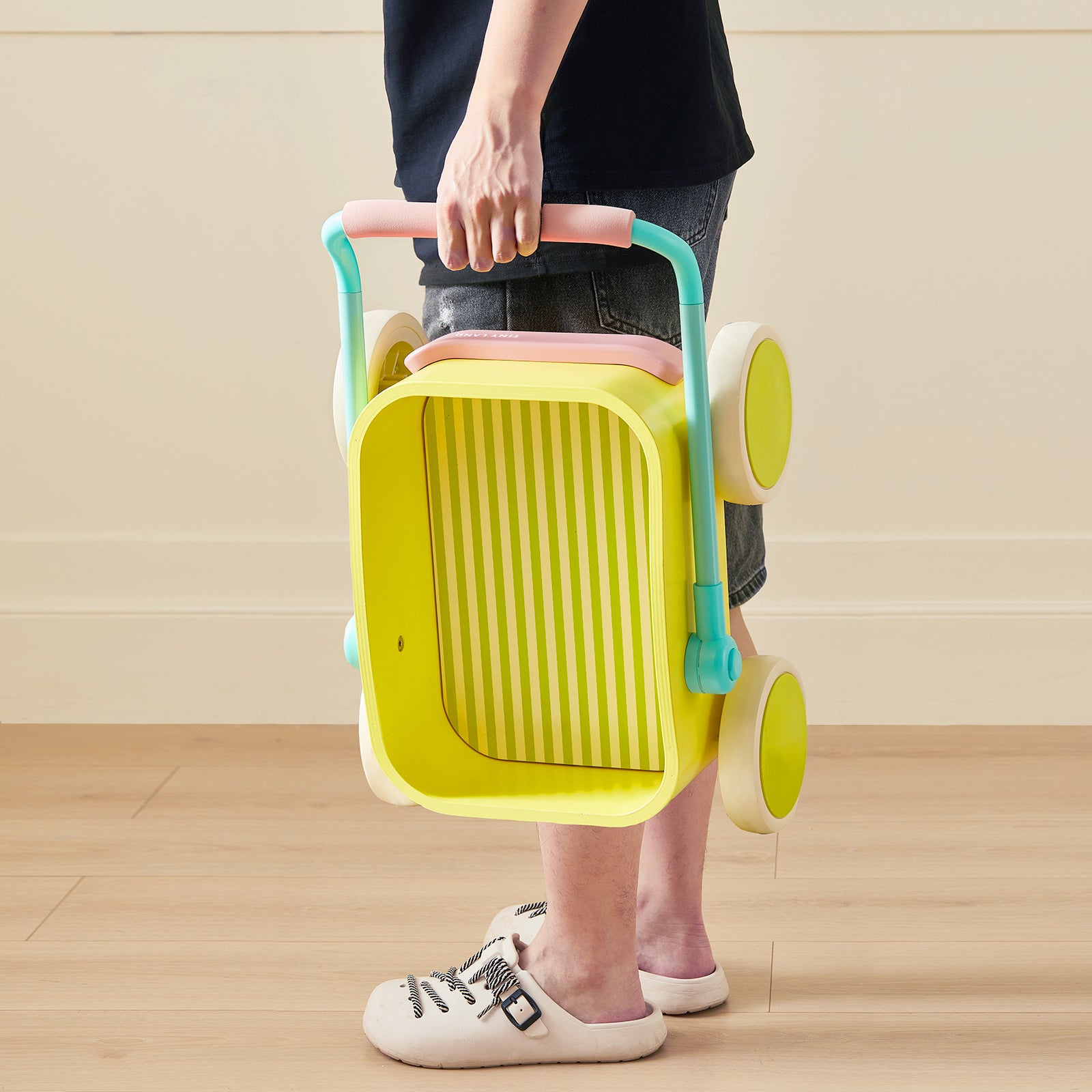 A person in shorts and slippers holds the Tiny Land® 3-in-1 Wooden Baby Walker Wagon - Spring Color by Tiny Land, featuring a colorful design, on a wooden floor against a cream wall.