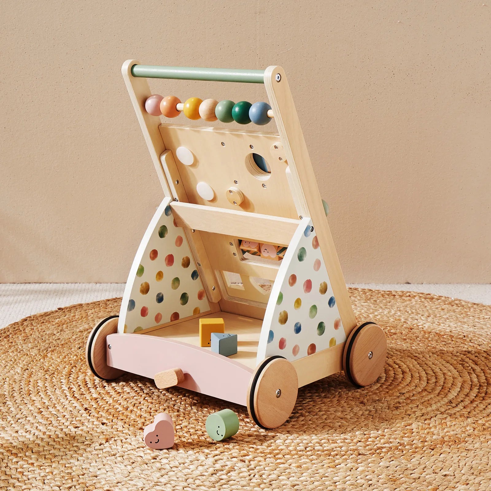The Tiny Land Complete Development Playset — Baby Walker + Activity Cube features a wooden baby walker with colorful beads, a shape sorter, and pastel polka dot sides, placed on a round woven mat to provide an engaging way to support fine motor skill development. Several wooden blocks are scattered nearby, creating an inviting, Montessori-inspired play area.