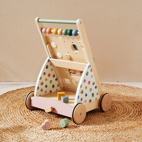 The Tiny Land Complete Development Playset — Baby Walker + Activity Cube features a wooden baby walker with colorful beads, a shape sorter, and pastel polka dot sides, placed on a round woven mat to provide an engaging way to support fine motor skill development. Several wooden blocks are scattered nearby, creating an inviting, Montessori-inspired play area.