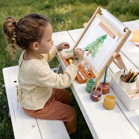 A young child sits at a white picnic table outdoors, painting a tree on the Tiny Land® Double-Sided Magnetic Tabletop Easel. Various paint jars and brushes from Tiny Land are arranged beside her on the table, creating a vibrant art station.|Oatmeal