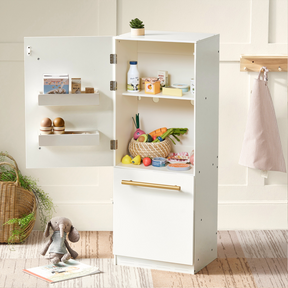A children’s play kitchen fridge with its door open shows Tiny Land® Organic Play Food Collection by Tiny Land Play Food, including realistic fruits and eggs. A stuffed rabbit sits nearby, and a striped apron hangs on the wall behind.