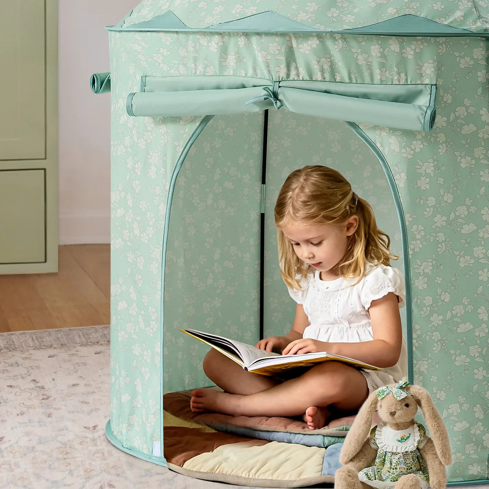 A young girl in a white dress reads inside the Tiny Land® Tiny Sprout Castle Tent by Tiny Land, sitting cross-legged on a quilted mat with a stuffed bunny nearby—a cozy and peaceful indoor scene.|Green