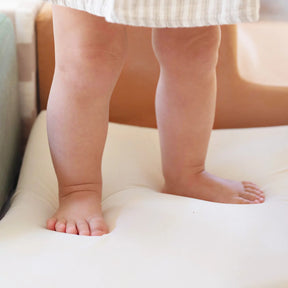 A young child stands barefoot on a soft Tiny Land® PlayNest Climbing Block by Tiny Land, with only their legs and feet visible beneath a striped garment.