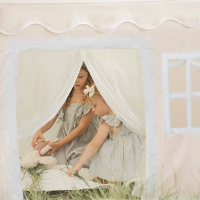 Two young girls in matching dresses play in the Tiny Land® Playhouse Tent – Natural Cream by Tiny Land, with one holding a stuffed animal. They sit on a white cushion as soft light fills this cozy indoor tent scene.|white