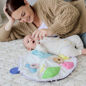 A woman in a beige sweater offers a toy from the Tiny Land® Montessori Toys for Newborns (0–3 Months) set to a smiling baby in a white onesie, who eagerly reaches out during tummy time on a patterned mat.