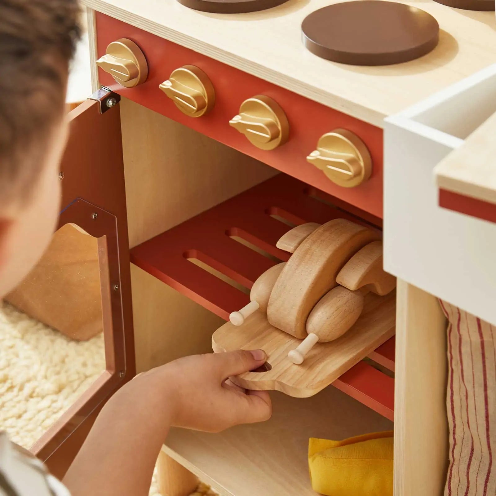 A child places a wooden toy chicken on a tray inside the Tiny Land® Mediterranean Play Kitchen oven, part of Tiny Land's wooden set featuring red and brown knobs|Mediterranean