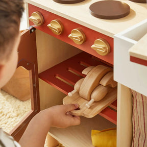 A child places a wooden toy chicken on a tray inside the Tiny Land® Mediterranean Play Kitchen oven, part of Tiny Land's wooden set featuring red and brown knobs|Mediterranean