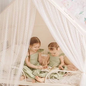 Two young girls in matching green dresses sit on a cozy Tiny Land® Chef’s Corner Playset – Cream Playhouse + Cream Kitchen Set, reading a book. The space has floral accents, soft lighting, and creates a warm, gentle playroom atmosphere.|mesh