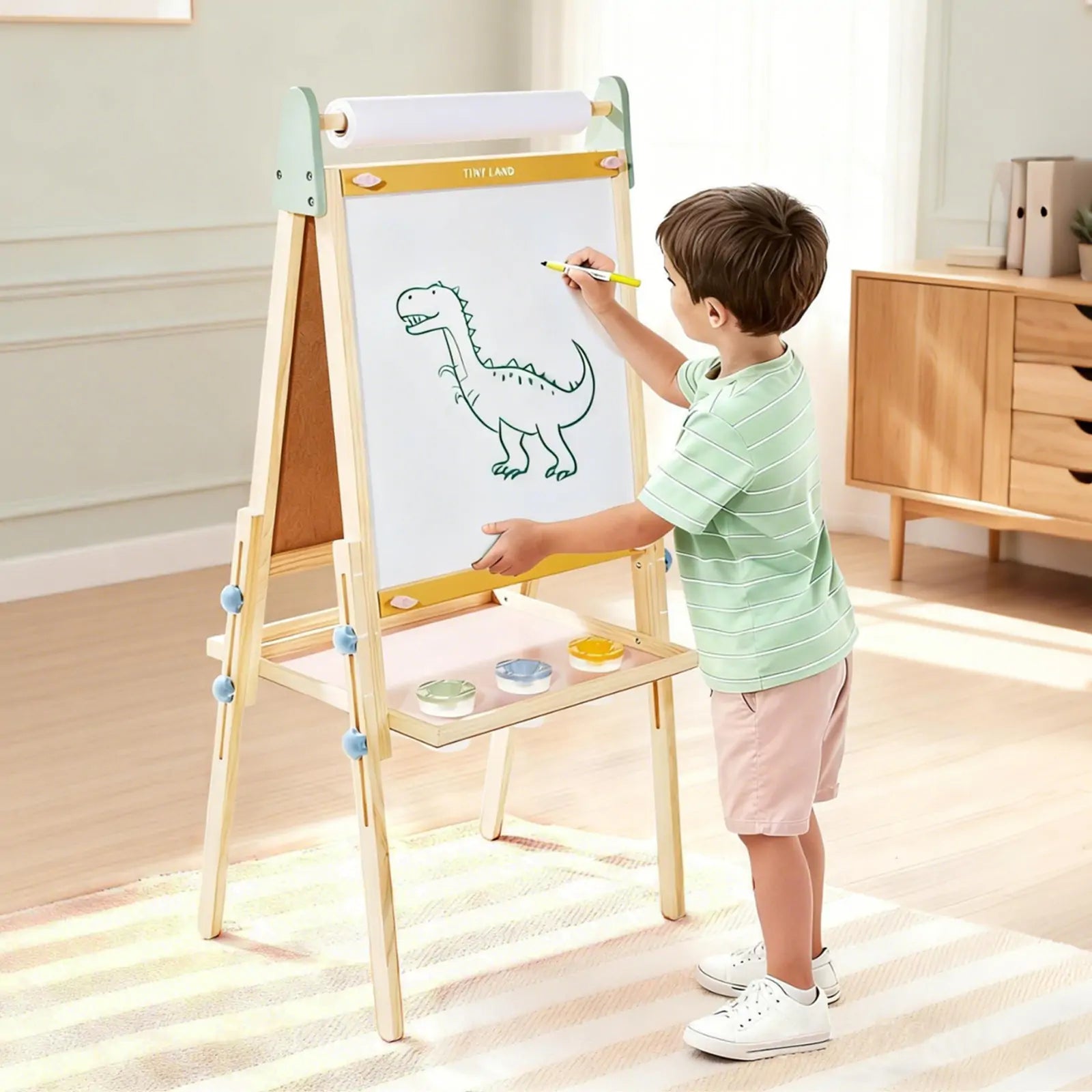 A young boy uses the Tiny Land® 3-in-1 Art Easel for Kids - Morandi, drawing a dinosaur on white paper with a yellow pencil in a bright, tidy room. Three paint pots rest on the shelf below the easel from Tiny Land|green