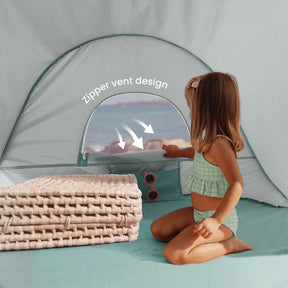 A young girl in a green swimsuit sits inside the Tiny Land® One-Stop Beach Toy & Tent Set – Sea Blue, peering through its zipper vent window as she enjoys the beach view next to two woven mats.|Sea Blue