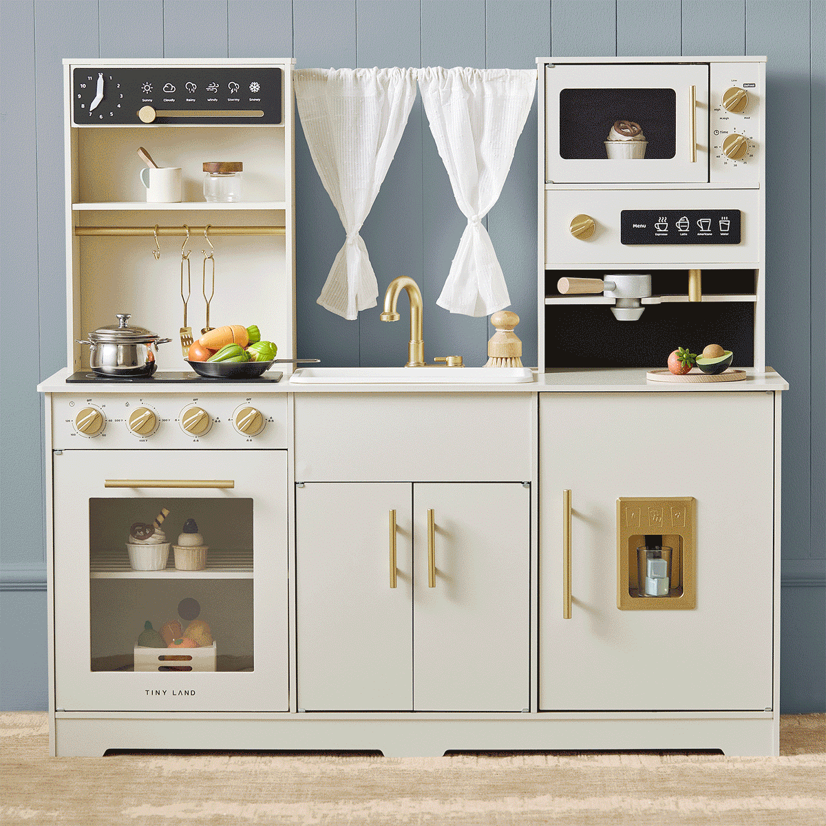 The Tiny Land® Play Kitchen with Running Water – Iconic Aqua Kitchen in Cream features a sink, stove, oven, microwave, gold handles, curtains above the sink, and includes play utensils and vegetables from Tiny Land.