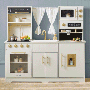 The Tiny Land® Iconic Water Play Kitchen & Premium Wooden Refrigerator by Tiny Land INC includes an oven, stove, sink with faucet, microwave, hanging utensils, play food, and a white curtain over a window—all showcased in a chic white and gold color scheme against a gray wall. This charming set is completed with a wooden cabinet refrigerator to inspire hours of imaginative pretend play for children.
