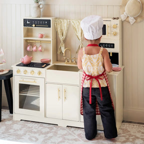 A child in a chef’s hat and apron plays at the Tiny Land® Iconic Wooden Play Kitchen—a cream cooking set with stove, oven, sink, microwave, and hanging cups in light colors with gold accents—pretending to cook.