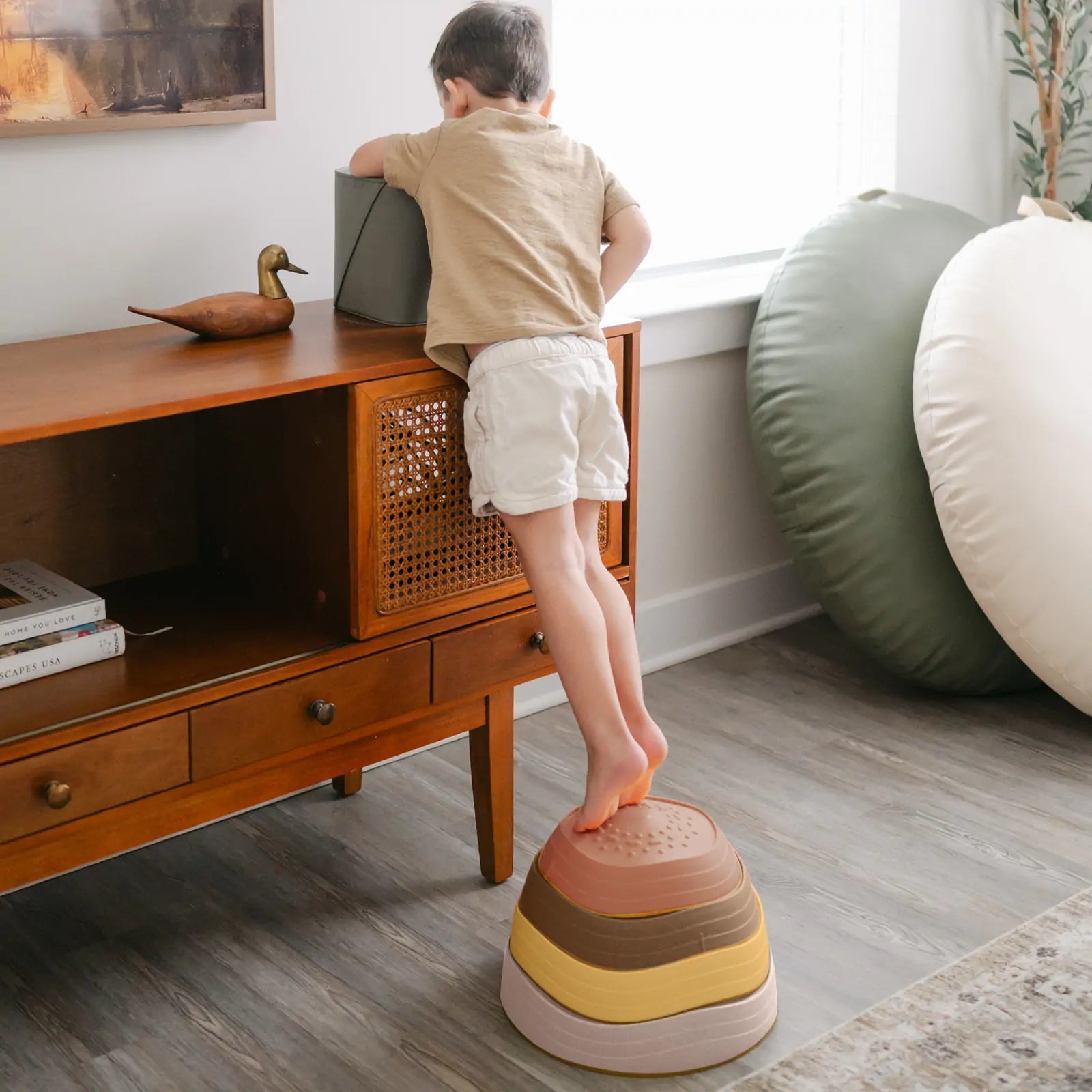 A young child uses Tiny Land® Stepping Stones - Sunset Hues by Tiny Land to build balance and coordination while reaching for a box atop a wooden cabinet in a bright, modern living room.|Sunset Hues