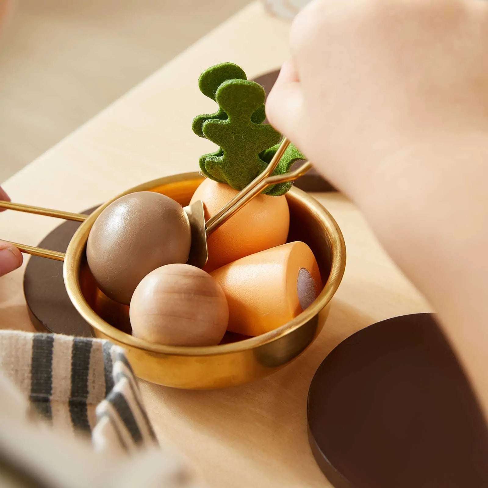 A person uses tongs to pick up a green felt vegetable from a brass bowl of wooden veggies on the Tiny Land® Mediterranean Play Kitchen by Tiny Land|Mediterranean