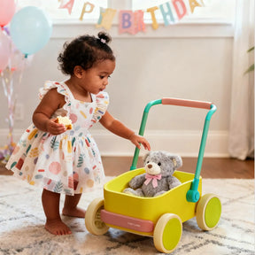 A toddler in a colorful dress enjoys a cupcake while playing with the Tiny Land® 3-in-1 Wooden Baby Walker Wagon - Spring Color, carrying a gray teddy bear. Birthday decorations and balloons are seen in the background.