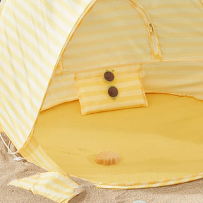 The Tiny Land® One-Stop Beach Toy & Tent Set in pastel yellow stripes sits on the sand, with a matching pillow holding round sunglasses and a seashell beneath its UV-protective shade—perfect for kids' beach fun.|Pastel Yellow Stripes