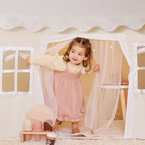 A young girl smiles in the doorway of the Tiny Land® Playhouse Tent, a natural cream indoor tent for kids by Tiny Land, decorated with soft curtains and a small table inside|white
