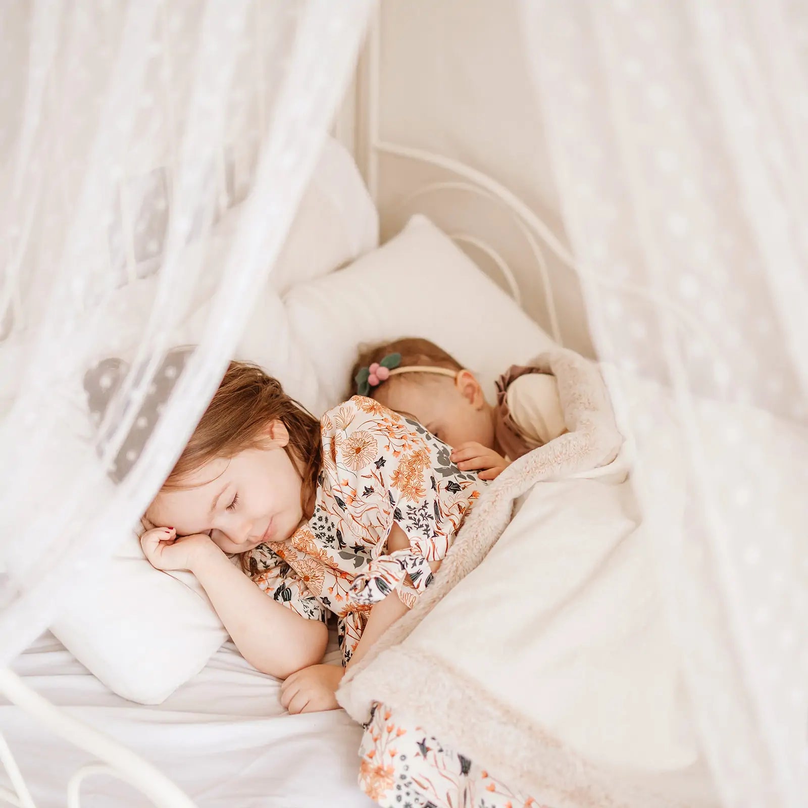 Two young children in floral pajamas sleep peacefully under a white blanket, surrounded by soft pillows and sheer polka-dot curtains—perfect after a day enjoying their Tiny Land® Chef’s Corner Playset – Cream Playhouse + Cream Kitchen Set.|mesh