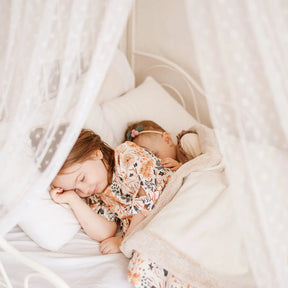 Two young children in floral pajamas sleep peacefully under a white blanket, surrounded by soft pillows and sheer polka-dot curtains—perfect after a day enjoying their Tiny Land® Chef’s Corner Playset – Cream Playhouse + Cream Kitchen Set.|mesh