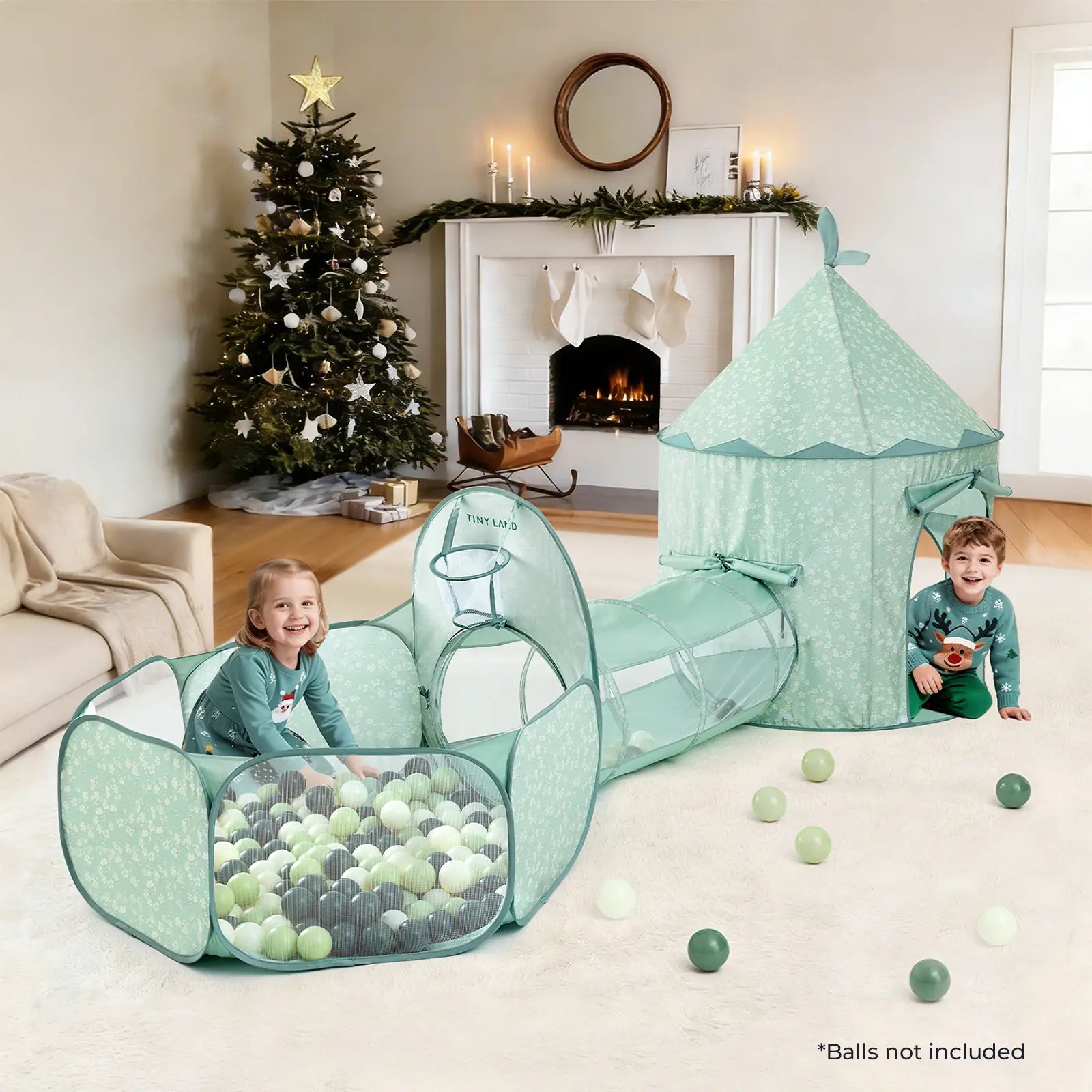 Two young children play in a Tiny Land® Compact Adventure 3-in-1 Play Tunnel With no Balls - Green, which features a cozy tent and tunnel. The living room is decorated for Christmas with a tree and fireplace, and some plastic balls are scattered on the carpet.|green