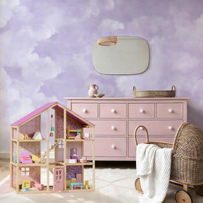 A pastel-themed child’s room featuring a pink dresser, Tiny Land® Sweetwood Unicorn Dollhouse & Wooden Dollhouse Dolls, a wicker bassinet with a white blanket, and a mirror on a cloud-painted purple and white wall.|Unicorn