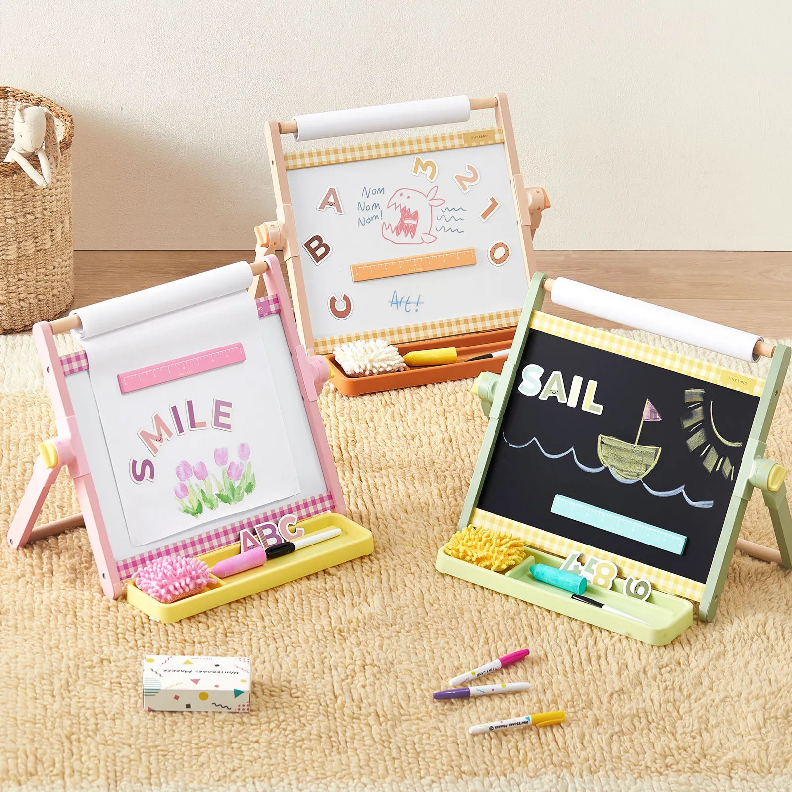 Three Tiny Land® Double-Sided Magnetic Tabletop Easels by Tiny Land, including an adjustable kids’ easel with a magnetic whiteboard, display colorful drawings and words. Each art station features storage trays holding chalk, erasers, and crayons—all set on a beige carpet in a cozy playroom setting.