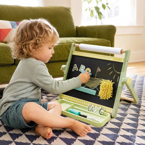 A young child sits on a rug, drawing with chalk on the Tiny Land® Double-Sided Magnetic Tabletop Easel from Tiny Land, which also features a magnetic whiteboard displaying block letters “ABC.” A green sofa and a window are in the background, while nearby art supplies help create a cozy art station.|Green